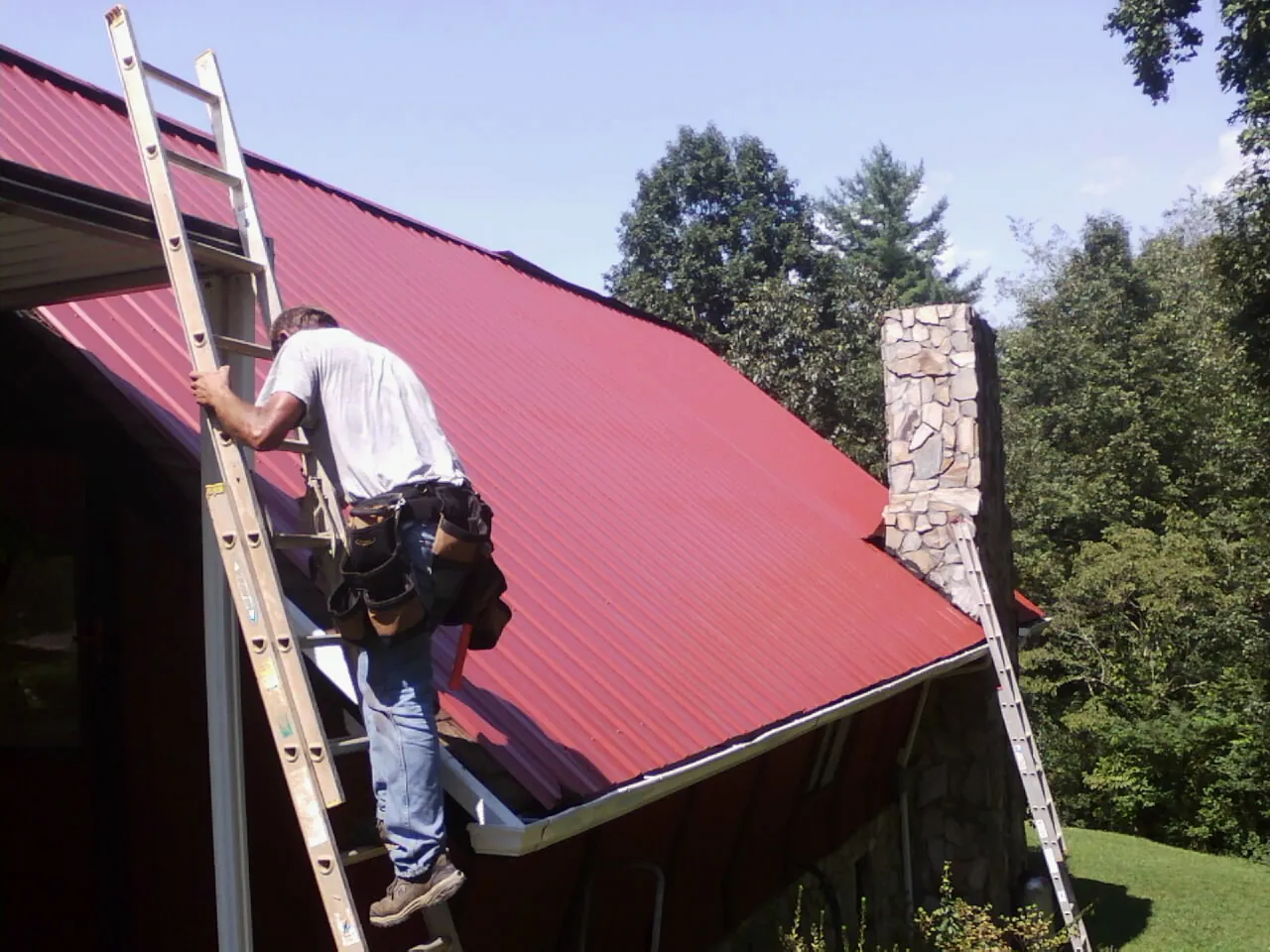 Roofer on a ladder beside a red metal roof and stone chimney — Soffit & Fascia Repair by Roofing Brothers in South Fulton